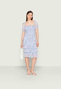 Vestido floral azul claro com mangas bufantes e design em camadas com babados. Apresenta um decote quadrado e é combinado com sandálias brancas de tiras.