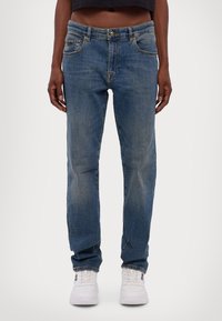 Kék farmernadrág, egyenes szabású dizájn, világos mosás, elhalványult részletekkel, öt zsebbel és fém kiegészítőkkel, fehér sportcipővel párosítva.