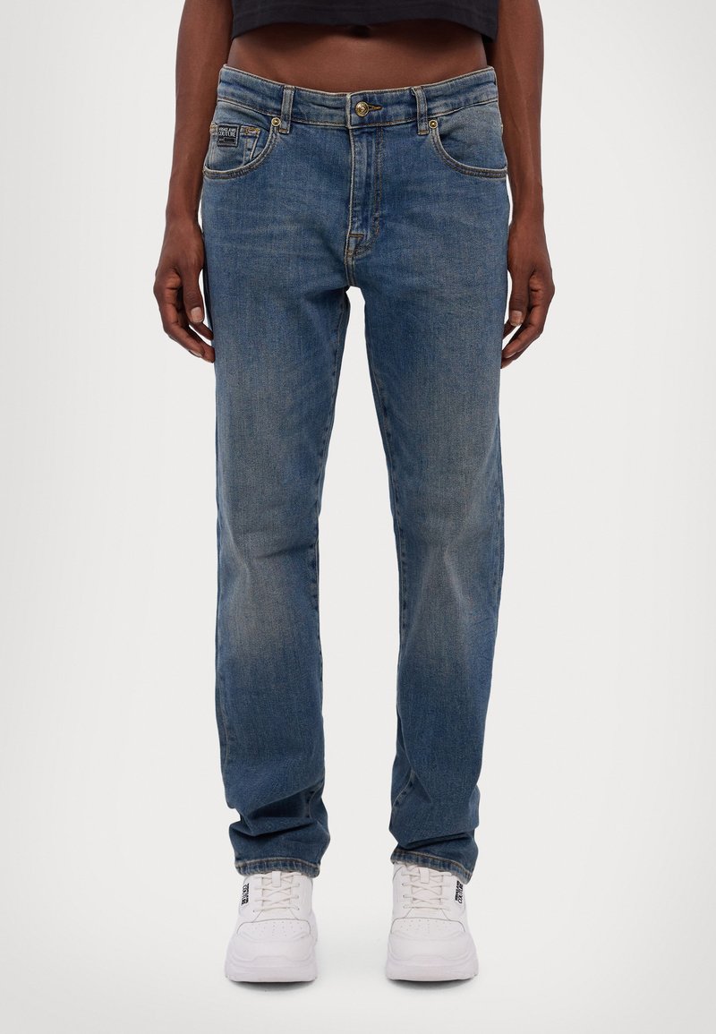 Kék farmernadrág, egyenes szabású dizájn, világos mosás, elhalványult részletekkel, öt zsebbel és fém kiegészítőkkel, fehér sportcipővel párosítva.