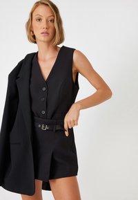 Mono negro sin mangas con escote en forma de V, cierre de botones en el frente y cintura ceñida con cinturón. Tejido texturizado y corte entallado. Acompañado de una chaqueta negra.