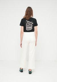 Svart t-skjorte med kakegrafikk og teksten "CONVERSE", kombinert med hvite vide bukser og mørkegrønne joggesko.