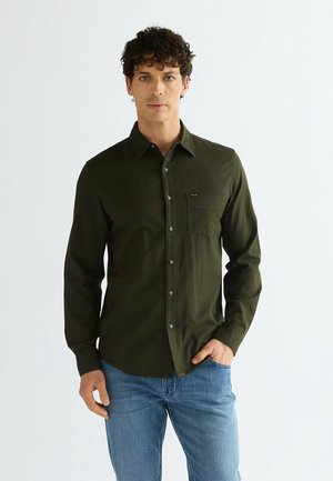 Mann mit lockigem dunklem Haar, der ein dunkelgrünes Hemd mit Knöpfen und blaue Jeans trägt, steht mit einer Hand in der Tasche.