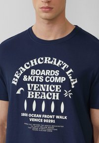Donkerblauwe katoenen t-shirt met witte grafische tekst, inclusief logo's en locatiegegevens, zoals "Venice Beach" en verschillende golficonen.