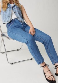 Blå denimhög midja jeans i kombination med en ljusblå denimväst med klassisk design, sittande på en transparent stol. Svarta remsandaler.