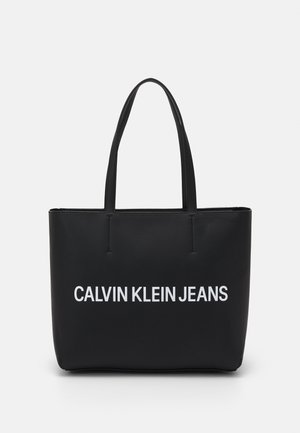 Sort tote-taske med to håndtag, med hvid "CALVIN KLEIN JEANS" tekst centreret på fronten mod en enkel baggrund.