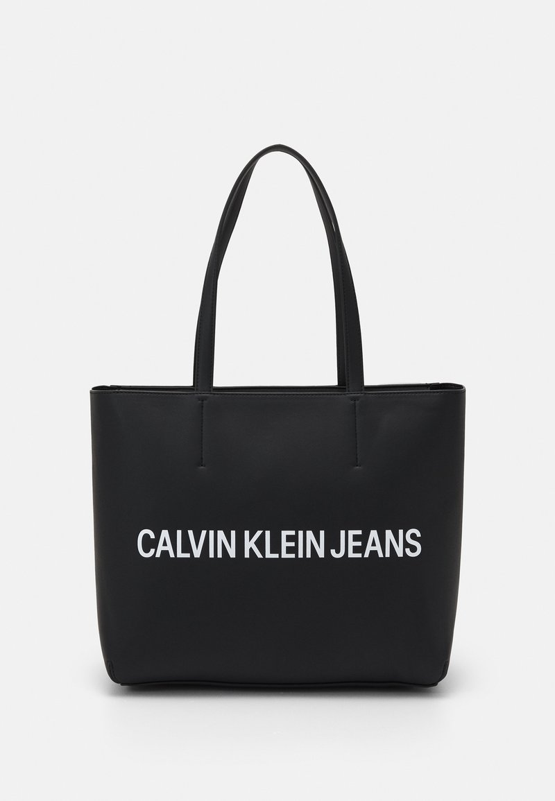 Sac fourre-tout noir avec deux poignées, avec le texte blanc « CALVIN KLEIN JEANS » centré à l’avant sur un fond uni.