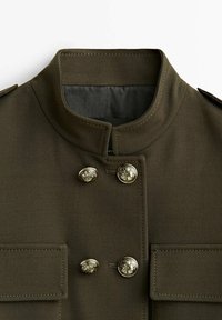 Veste de style militaire vert olive avec col montant, deux poches à rabat et quatre boutons métalliques décoratifs avec blasons embossés.
