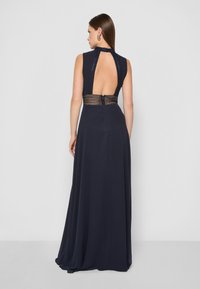 Námornícky modré večerné šaty s vysokým výstrihom, odhaleným chrbtom a zložitými čipkovými detailmi v oblasti pásu; dlhá splývavá sukňa sa vlečie po podlahe.