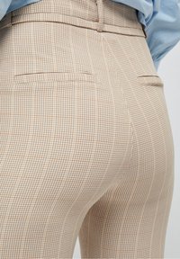 Kárované kalhoty v béžové a hnědé barvě s přiléhavým střihem, obsahující dvě zadní kapsy a hladkou texturu.