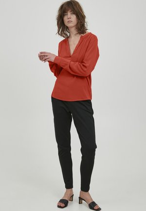 Rustfarbene Bluse mit V-Ausschnitt, langen Ärmeln und einem lockeren Schnitt. Kombiniert mit schwarzen, schmal zulaufenden Hosen und schwarzen Slipper-Sandalen.