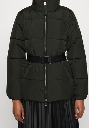 Sort quiltet dunjakke med høj krave, lynlås foran, bælte i taljen, lynlåslommer, båret over en sort plisseret nederdel.