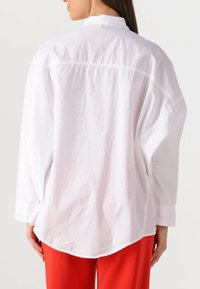 Weißes Langarmhemd mit abgerundetem Saum, das ein Detail mit Vorderstoß und ein Etikett am Kragen aufweist. Kombiniert mit roten Hosen.