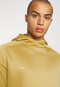 Huvtröja i ljusgul färg, tillverkad av slätt tyg. Har en logotyp på bröstet och en avslappnad passform med en strukturerad huva.