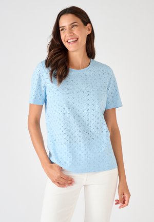 Donna sorridente che indossa una maglietta azzurro chiaro a maniche corte con occhielli e pantaloni bianchi, in piedi su uno sfondo semplice e chiaro.