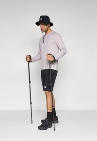 Gaiši violeta garām piedurknēm krekls, melnas šortas, melnas pārgājienu zābaki un melna buĻīša cepure. Tur rokās pārgājienu nūjas, stāv taisni.