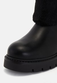 Botină neagră din piele, cu vârful rotunjit, ornament din blană texturată și o talpă robustă, zimțată, de culoare neagră, care are cusături decorative.