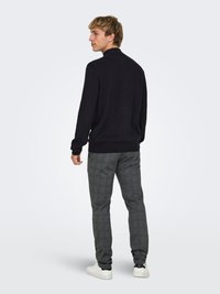 Maglione nero a maglia, pantaloni grigi a quadri e sneakers bianche. Il maglione ha una vestibilità classica; i pantaloni presentano un motivo strutturato e gambe affusolate.