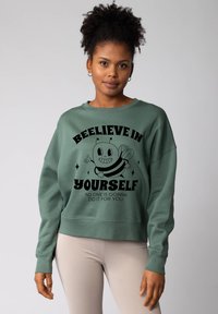 Groene katoenen sweatshirt met een zwart ontwerp van een glimwesp en de tekst "GELIEF IN JEZELF" met een geribbelde textuur en een losse pasvorm.