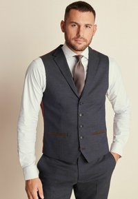 Chaleco de mezcla de lana navy con cinco botones, acentos en marrón, bolsillos laterales y un panel rojo en contraste. Lleva puesto sobre una camisa clara a rayas y una corbata.