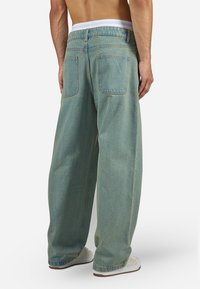 Mand iført løstsiddende, falmede blå jeans og hvide sneakers, vist fra ryggen, stående mod en ensfarvet baggrund.