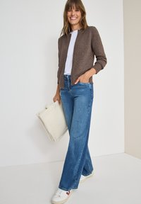 Brun stickad cardigan, vit T-shirt, blå vida jeans, håller en krämfärgad texturerad tote-väska, paired med vita sneakers med röda detaljer.