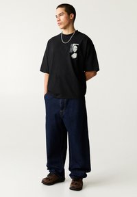 Schwarzes T-Shirt mit kurzen Ärmeln und grafischem Druck. Kombiniert mit lockeren, dunkelblauen Jeans und braunen Sneakern, die über ein strukturiertes Material und kontrastierende Nähte verfügen.