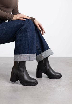 Stivali in pelle nera alla caviglia con tacco grosso e suola testurizzata, dotati di una cerniera laterale e abbinati a jeans di denim scuro arrotolati ai polsi.