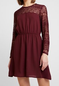 Robe bordeaux en dentelle et tissu, avec des manches longues en dentelle, une taille élastique et une jupe évasée.