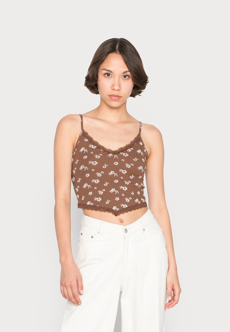 Top cropped com padrão floral castanho e acabamento em renda, apresentando alças finas de espaguete e textura ribada. Combinado com calças brancas de perna larga.