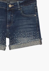 Shorts de mezclilla en azul oscuro, con un dobladillo deshilachado y decorados con tachuelas iridiscentes en la parte inferior para un detalle adicional.