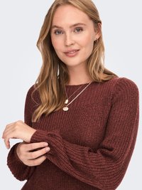 Maglione a maglia bordeaux con texture a coste, maniche lunghe e vestibilità rilassata. Abbinato a delicate collane d'oro e una posizione delle mani morbida.