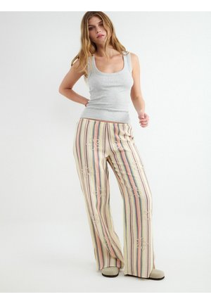 Kobieta stojąca, ubrana w jasnoszary top bez rękawów oraz luźne, wielopaskowe spodnie w pastelowych kolorach z lekkim przetarciem.