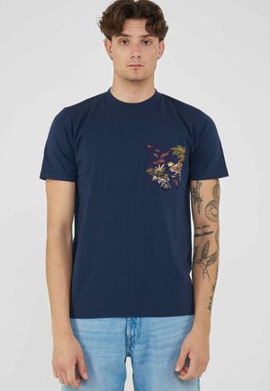 Jonge man met krullend haar die een marineblauw t-shirt draagt met een kleurrijk tropisch patroon op de borstzak en lichtblauwe spijkerbroek, toont armtatoeages.