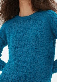 Tealfärgad stickad tröja med ett strukturerat mönster och kabelstickade ärmar. Rund halsringning, lång ärm och ribbad fåll.