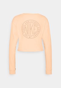 Peach long-sleeve cropped shirt with a round logo on the back featuring the word "NIKE" and text in a circular design. Smooth fabric texture.
