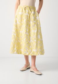 Jupe midi à motifs floraux jaunes en tissu texturé, dotée d'une taille élastique froncée et de poches latérales. Associée à des chaussures beiges.