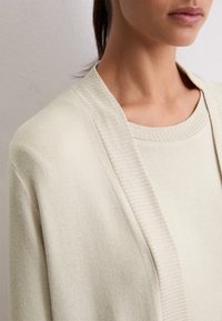 Donna che indossa un maglione lavorato a maglia beige chiaro con colletto a costine e un cardigan abbinato, mostrata dalle spalle alla bocca su uno sfondo di parete chiara.