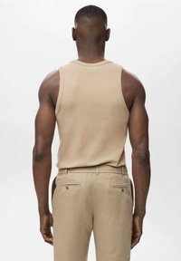 Beige ærmeløs tanktop, ensfarvet design, båret med lyse beige bukser med baglommer. Glat stof; tætsiddende silhuet.