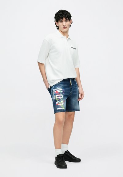 Polo blanc, short en jean bleu avec imprimé graphique coloré, baskets noires et chaussettes blanches montantes. Tenue décontractée avec une coupe confortable.