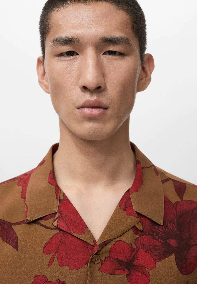 Camicia a maniche corte marrone con motivi floreali rossi di fiori e foglie, con colletto classico e chiusura con bottoni. Tessuto dalla texture liscia.
