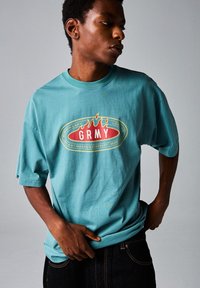 Camiseta de algodón turquesa con corte oversized, que presenta un diseño gráfico central en rojo y amarillo que incluye la palabra "GRMY" y acentos de llamas.