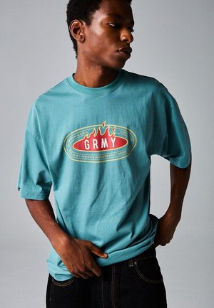 Camiseta de algodón turquesa con corte oversized, que presenta un diseño gráfico central en rojo y amarillo que incluye la palabra "GRMY" y acentos de llamas.
