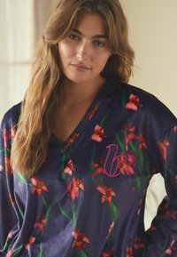 Tumesinine lilleline pluus erksate roosa ja rohelise mustriga, millel on V-kaelus ning rinnal tikkitud kursiivne "b" kujundus.