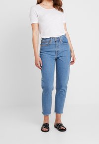 Camiseta blanca ligera combinada con jeans azules de cintura alta, con pierna recta, dobladillo desgastado y sandalias negras con detalles de tachuelas.