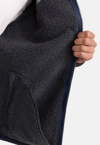Das Innenfutter der Fleecejacke zeigt einen weichen, strukturierten dunkelgrauen Stoff mit einem Reißverschlussrand und einer Tasche auf der linken Seite.