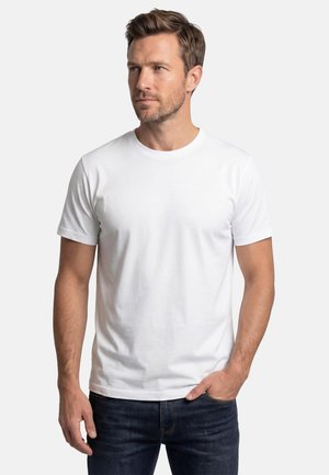 Mann mit hellbraunen Haaren, der ein schlichtes weißes T-Shirt mit kurzen Ärmeln und dunkelblaue Jeans trägt und mit einer Hand in der Tasche steht.