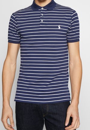 Mann trägt ein navyblaues Poloshirt mit weißen horizontalen Streifen und einem bestickten Pferdelogo, kombiniert mit dunklen Jeans.