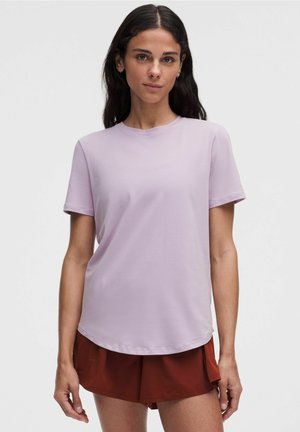 Kobieta o długich ciemnych włosach, ubrana w jasnofioletowy t-shirt z krótkim rękawem i szorty w kolorze rdzy, stojąca na jednolitym tle.