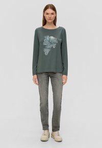Donkergroene top met lange mouwen en een grafische bloemenprint in blauw en wit, gecombineerd met grijze jeans en crèmekleurige sneakers. Gladde stof, losse pasvorm.