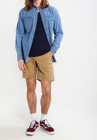 Chemise en denim bleu clair, débardeur bleu marine, short beige et baskets bordeaux avec semelles et lacets blancs. Détails des boutons visibles sur la chemise.